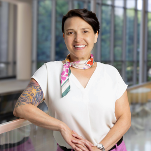 Portrait of Öge Gözütok Moore wearing a colorful scarf and short sleeved blouse. She leans on the stairwell railing in the modern glass Louis A. Simpson and Kimberly K. Querrey Biomedical Research Center building. She graduated with a master's degree in regulatory compliance in 2025.