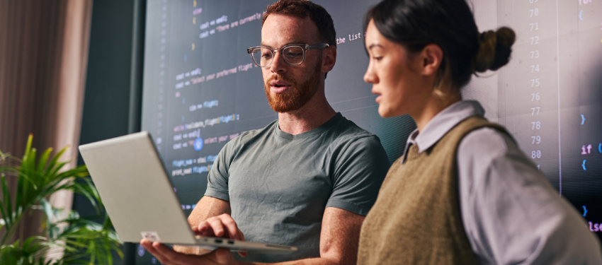 Two information systems professionals review a presentation on a laptop. A man with glasses and t-shirt holds the laptop while his colleague, a woman in a sweater vest over a collared shirt, looks over his shoulder.