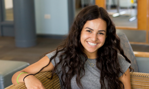 Gabrielle has long curly hair and wears a casual gray t-shirt. She is smiling, lounging on a couch in a college common room.