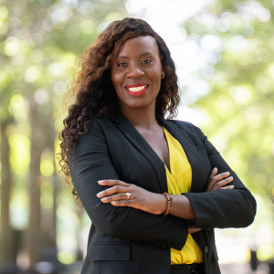 Rose Kargbo in a park at Iowa, with green trees lining the street on a beautiful sunny summer day. She wears a professional suit, bright yellow top, and has her arms crossed in a professional pose. Rose graduated with an MS in Global Health from Northwestern SPS in 2023.
